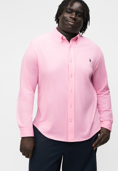 Mann med mørk hud iført en lys rosa skjorte med lange ermer og mørke bukser, stående mot en enkel bakgrunn.