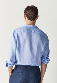 Vaaleansininen pellavapaita, rento istuvuus, pitkähihaiset hihat käärittyinä ja kaulus ilman käännettä. Takakuvassa näkyy minimaalinen ompelu ja sileä pinta.