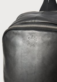 Zwarte leren rugzak met een gladde textuur, voorzien van een zilveren rits en een reliëflogo-detail. Schone lijnen en een minimalistisch ontwerp.
