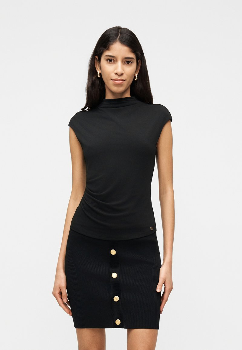 Γυναίκα με μακριά σκούρα μαλλιά, που φοράει μαύρο αμάνικο τοπ και μαύρη φούστα με μια κάθετη σειρά από πέντε χρυσά κουμπιά στο μπροστινό μέρος.