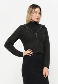Svart cropped zip-up hettegenser med et teksturert mønster, hette og lange ermer, båret over en tettsittende svart kjole. Glatt stoff.