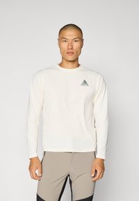Langarm-Cremeshirt aus leichtem Material mit einem kleinen türkisfarbenen dreieckigen Logo auf der linken Brust und flachen Nähten.