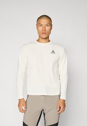 Lange mouw crème shirt gemaakt van lichte stof met een klein teal driehoekig logo op de linkerborst en platte naden.