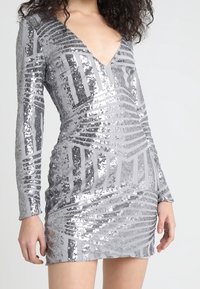 Robe à sequins argentés avec des manches longues, décolleté en V profond et motif en bandes géométriques. Tissu près du corps avec un éclat texturé partout.