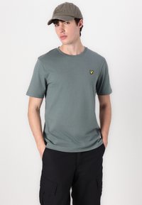 Camiseta de algodón verde con mangas cortas y un escote redondo, que presenta un pequeño logo amarillo bordado en el pecho. Corte casual.