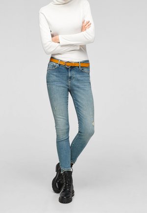 Personne en jean skinny bleu clair, col roulé blanc, ceinture orange et bottes noires à lacets, debout les bras croisés.