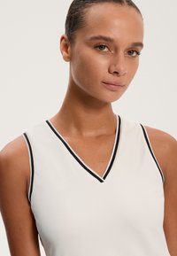 Ärmlös vit topp med V-ringning, med en kontrasterande svart kant längs halsringningen och ärmhålen. Slät tygtextur.