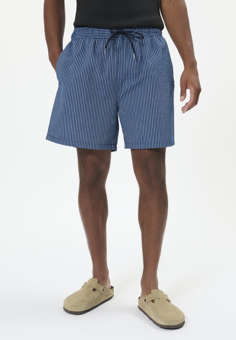 Pantaloni corti a righe blu e bianche con cintura elastica e lacci, dotati di tasche laterali. Indossati con scarpe beige senza lacci.