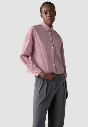 Woman wearing a red and white striped button-up shirt with a chest pocket and high-waisted gray pleated trousers, standing against a plain background.