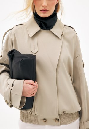 Kobieta w beżowej, oversize kurtce i czarnym golfie trzyma dużą złożoną czarną skórzaną kopertówkę na białym tle.