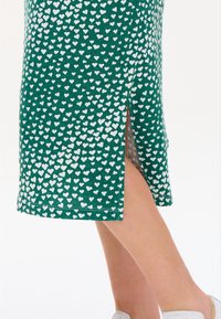 Green dress with white heart pattern, knee-length, featuring side slit and soft fabric texture. Styled with light-colored shoes.