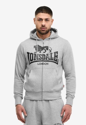 Uomo con felpa con cappuccio grigia Lonsdale London con zip e pantaloni della tuta abbinati, in piedi su sfondo bianco semplice.