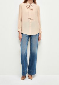 Blusa rosa chiaro a maniche lunghe con fiocco al collo, abbinata a jeans azzurri a gamba larga con orlo sfilacciato e scarpe a punta marroni.