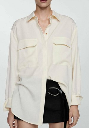 Camicia button-up beige chiaro con due grandi tasche, vestibilità affusolata, maniche lunghe arrotolate ai polsi, bottoni a contrasto e texture liscia.