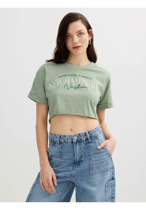 Joven mujer con cabello largo y oscuro, que lleva pendientes de aro dorados, una camiseta corta verde claro con el texto "New Hampshire" y vaqueros azules de talle alto.