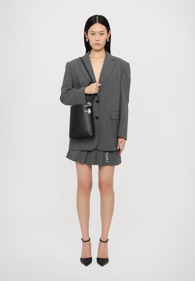 Blazer gri cu dungi, cu umeri mari, o fustă mini plisată asortată cu textul "SHORT", o geantă neagră tip bucket și pantofi cu toc ascuțit.