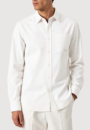 Uomo che indossa una camicia bianca a manica lunga con taschino sul petto e logo ricamato, abbinata a pantaloni chiari.