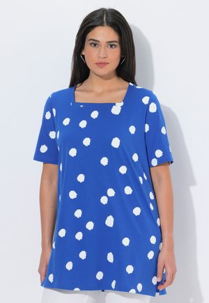 Donna che indossa una maglietta blu a maniche corte con pois bianchi irregolari e scollo quadrato, su uno sfondo chiaro e uniforme.