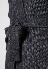 Mörkgrå stickad plaggdetalj med ett texturerat mönster, bundet skärp i midjan och en mjuk, vävd framtoning.