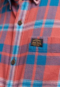 Red and blue plaid flannel shirt with a textured finish, featuring brown buttons and a black label with gold text.