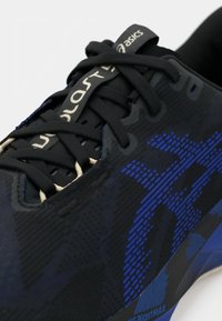 Baskets de sport noires avec des accents bleus, présentant un dessus texturé, un design à motifs et un laçage standard. Logo visible sur la languette.