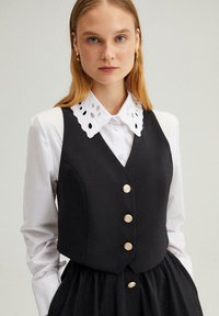 Svart ärmlös väst med fem guldknappar, bärs över en vit skjorta med broderie anglaise-krage, med en slät yta och figursydd design.