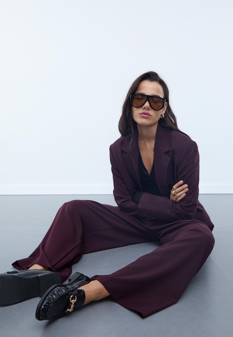 Bordová oversized sako, čierny top, veľké hnedé slnečné okuliare a čierne šľapky so zdobným reťazovým akcentom. Hladká tkanina, voľný strih.