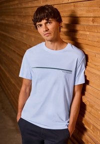 Un t-shirt en coton bleu clair présente un imprimé graphique horizontal vert foncé sur la poitrine. Le modèle est adossé à un mur en bois.