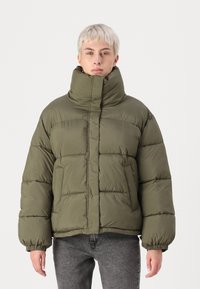 Olivovo zelená puffa bunda s vysokým golierom, štvorcovým strihom, elastickými manžetami a viditeľným prešívaním. Má hladký, matný povrch.