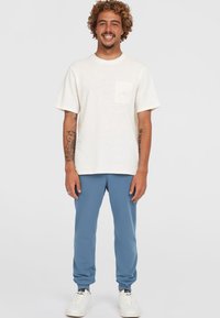 White short-sleeve t-shirt with a chest pocket, paired with blue sweatpants, worn with white sneakers. Casual outfit with a relaxed fit.