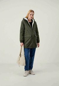 Γυναίκα με πράσινο μπουφάν με κουκούλα, μπλε τζιν, λευκά αθλητικά παπούτσια, κρατώντας μια μπεζ τσάντα με γέμισμα, που στέκεται μπροστά σε έναν απλό φωτεινό φόντο.