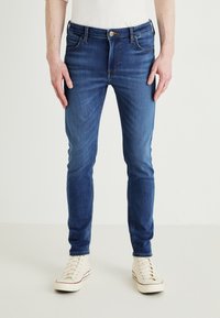 Εφαρμοστά τζιν παντελόνια σε σκούρο μπλε απόχρωση με λείο υφή, πέντε τσέπες και κουμπάκι. Συνδυασμένα με αθλητικά παπούτσια σε ανοιχτό χρώμα.