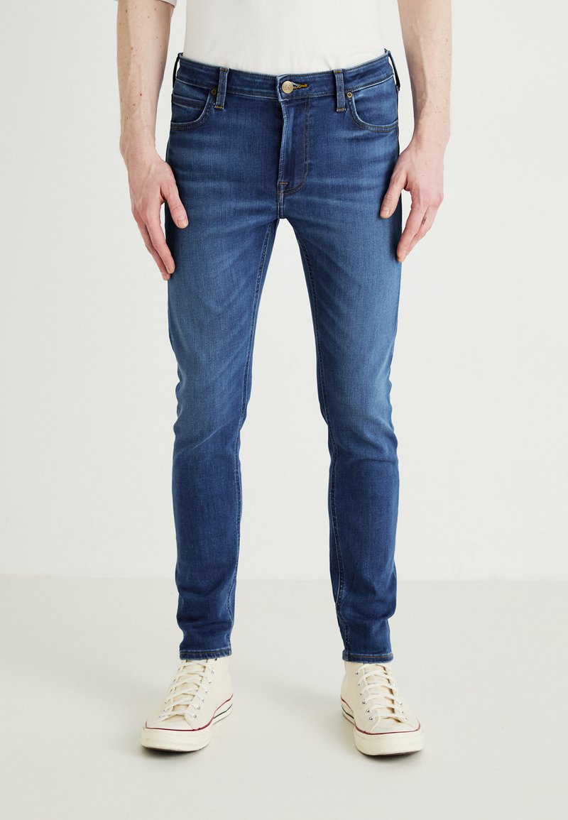 Εφαρμοστά τζιν παντελόνια σε σκούρο μπλε απόχρωση με λείο υφή, πέντε τσέπες και κουμπάκι. Συνδυασμένα με αθλητικά παπούτσια σε ανοιχτό χρώμα.