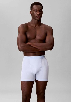 Muscular man standing with arms crossed, wearing white Calvin Klein boxer briefs against a plain gray background.