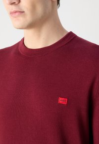Gros plan d'une personne portant un pull ras du cou bordeaux avec une petite étiquette rouge "HUGO" sur le côté gauche de la poitrine.