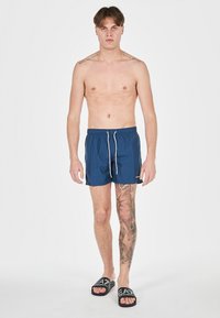 Bañadores azul marino con cordón; tejido ligero; el modelo lleva chanclas negras; presenta tatuajes en la pierna izquierda.
