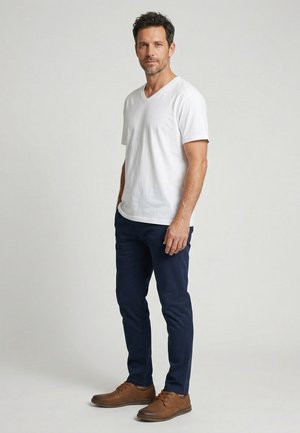 Mand stående iført en hvid kortærmet V-hals skjorte, marineblå bukser og brune lædersko mod en ensfarvet lys baggrund.