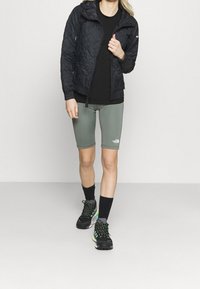 Černá prošívaná bunda s kapucí, černé tričko, světle šedé cyklistické krátké kalhoty a černé boty se zelenými akcenty. Černé ponožky dotvářejí outfit.