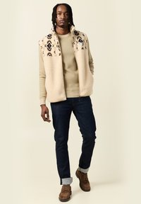 Gilet in pile beige con parte superiore decorata, maglione color beige, jeans in denim scuro e stivali marroni con risvolti.