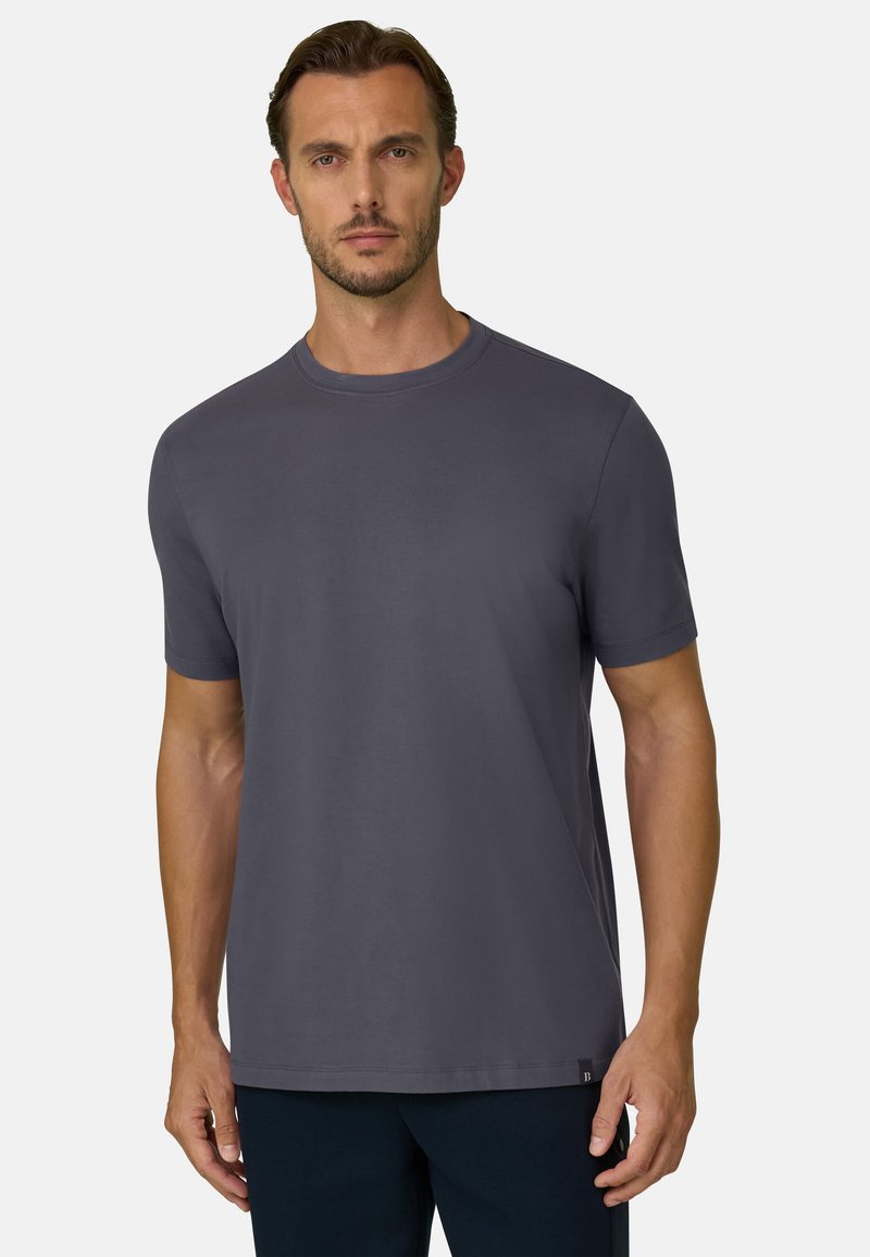 Homme portant un t-shirt uni gris foncé à manches courtes et col rond, debout devant un fond clair.