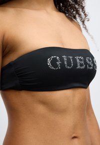Μαύρο τοπ bandeau από λείο ύφασμα, με το λογότυπο "GUESS" σε λαμπερές στρας. Ο σχεδιασμός διαθέτει ραφές στις πλευρές για επιπλέον σχήμα.