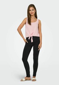Lichtroze mouwloze top met een geknoopte voorkant, gecombineerd met aansluitende zwarte jeans en platte witte sandalen. Soepele stoftextuur, casual ontwerp.