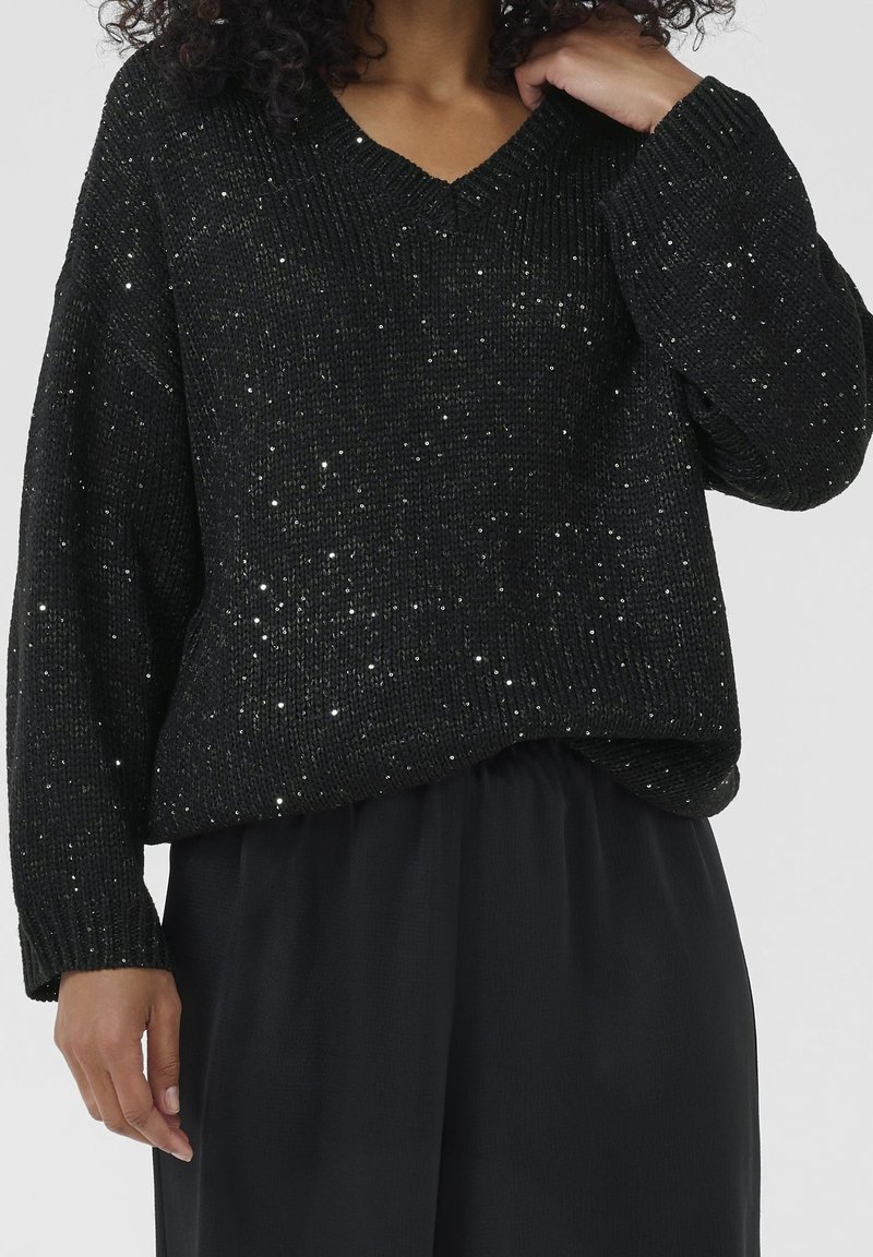 Femme portant un pull à col en V noir avec des détails scintillants argentés, associé à une jupe noire, la main touchant son cou.