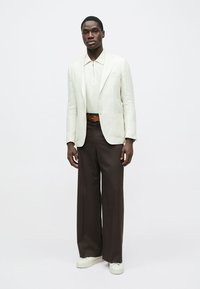 Blazer en lin de couleur claire porté sur un polo crème, associé à un pantalon brun foncé et des baskets blanches, mettant en avant une silhouette épurée et moderne.