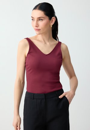 Vrouw met donker haar in een paardenstaart, die gouden oorbellen draagt, een mouwloze bordeaux rib top en een zwarte broek met één hand in de zak.