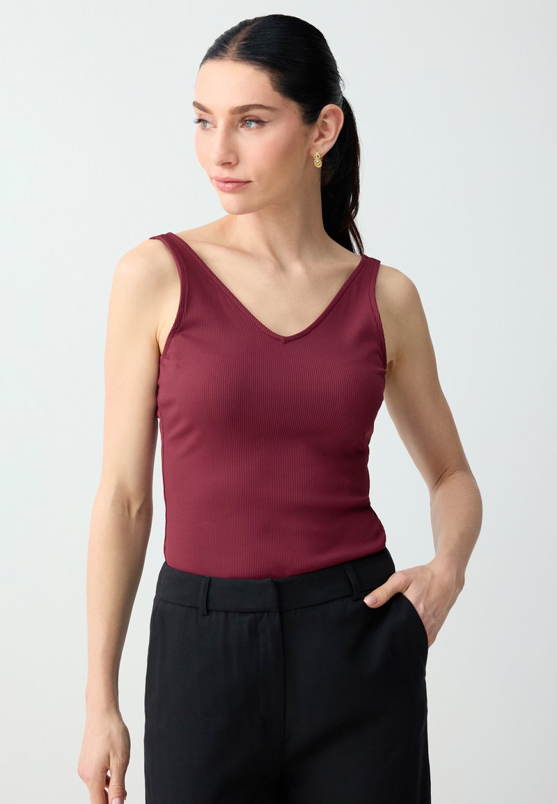 Femme aux cheveux foncés attachés en queue de cheval, portant des boucles d'oreilles en or, un top sans manches côtelé bordeaux et un pantalon noir, une main dans la poche.