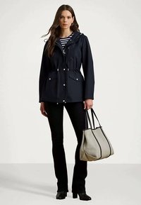 Ženska nosi temnomodru jaknu preko prugaste majice, crne hlače i crne čizme, drži veliku bež torbu s crnim obrubom.