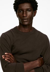 Braun dunkel langärmliges Shirt aus geripptem Strickstoff mit rundem Hals und eng anliegenden Bündchen. Das Modell hat die Arme verschränkt und einen neutralen Gesichtsausdruck.