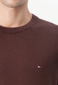 Barna kötött pulóver kerek nyakkal. Kicsi, hímzett logó található a mellkasán kék, fehér és piros színekben. Puha tapintású.