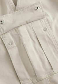 Tissu beige avec une texture lisse, comportant une poche avant, des boutons en métal et des coutures contrastées pour des détails structuraux.